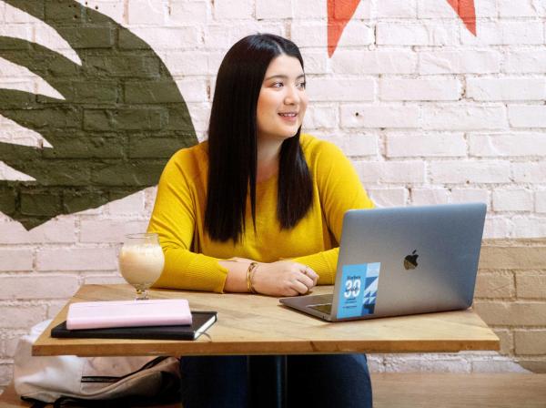 The image depicts an individual sitting at a wooden table with a laptop open in front of them. The laptop displays a blue graph or chart. To the left is a pink notebook, and in front of the person is a glass containing a creamy beverage. The person wears a yellow long-sleeved top. Behind them is an interior wall with a green artistic design or logo.