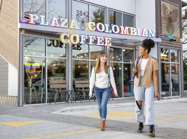 The image shows two women walking and chatting in front of "Plaza Colombian Coffee." One wears a white sweater, blue jeans, and brown boots, while the other has a light brown shirt, ripped jeans, and black boots. They are smiling, and the modern coffee shop has large windows and colorful signage. The interior appears cozy and inviting.