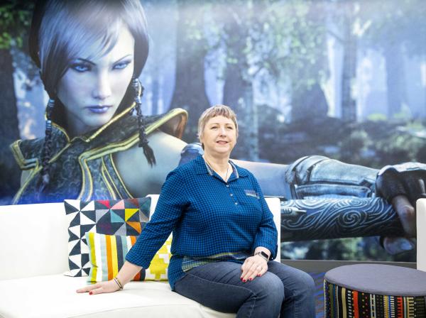 The image shows a woman sitting on a white couch in front of a large poster or screen displaying a character from a video game. The woman sitting on the couch is dressed in a blue checkered shirt and jeans, with short hair. She is smiling slightly and appears to be in a relaxed pose. There are colorful pillows on the couch, and a small round stool or table with a patterned cover is positioned next to the couch. The setting appears to be casual and modern, likely within a gaming or tech environment.