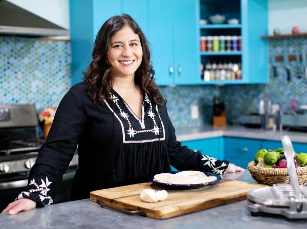 The image features a woman with long, dark hair, wearing a black blouse with white embroidery. She is standing in a bright kitchen with turquoise cabinets and a mosaic tile backsplash. She is smiling and appears to be preparing food, as there is a cutting board with dough and a plate with tortillas in front of her. A basket of vegetables and a tortilla press are also visible on the counter.