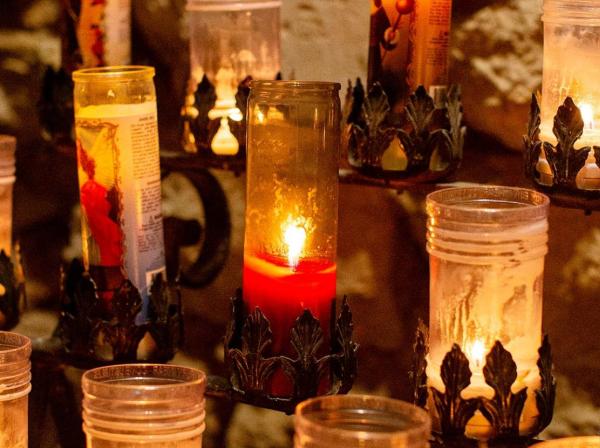 The image shows a collection of candles placed within glass holders. The candles vary in height and the amount of wax melted, suggesting they have been burning for different durations. Some candles are lit, casting a warm, inviting glow, while others remain unlit. The glass holders feature decorative bases, possibly made of metal with intricate designs. The background is blurred but appears to be a wall with patterns, likely reflections or shadows created by the candlelight.