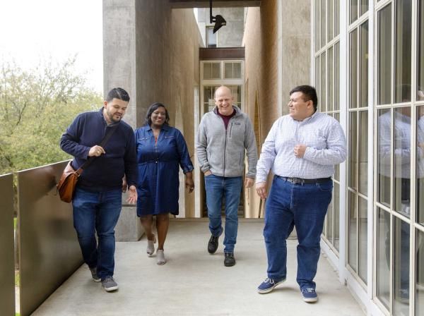 The image shows four people walking together along an outdoor corridor with large windows on the right and a railing on the left. The group consists of two men and two women, all casually dressed and engaged in conversation, creating a friendly and collegial atmosphere. The background includes greenery visible through the windows and an architectural setting with concrete and brick elements. The scene conveys a sense of collaboration and camaraderie.