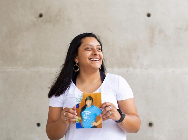 The image shows a young woman standing against a concrete wall. The woman is wearing a white t-shirt with some visible graphic designs on it. The person is holding a photograph that shows another individual wearing a blue t-shirt with some text or design on it. 