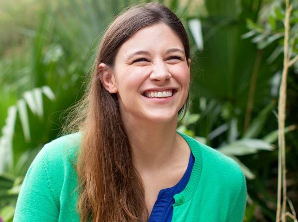 The image shows an individual standing in front of a lush background filled with green foliage. The person has long, light brown hair and is wearing a bright green top paired with a blue cardigan.