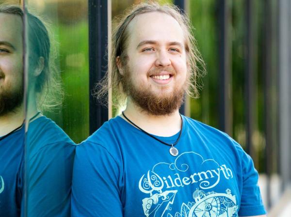 The image shows a bearded man with long hair, smiling while standing next to a reflective glass surface. He is wearing a blue T-shirt with the word "Wildermyth" and a detailed design on it. The reflection in the glass creates a mirrored effect, showing both his front and side profile. The background is blurred, suggesting an outdoor setting with natural light. He is also wearing a necklace with a circular pendant.