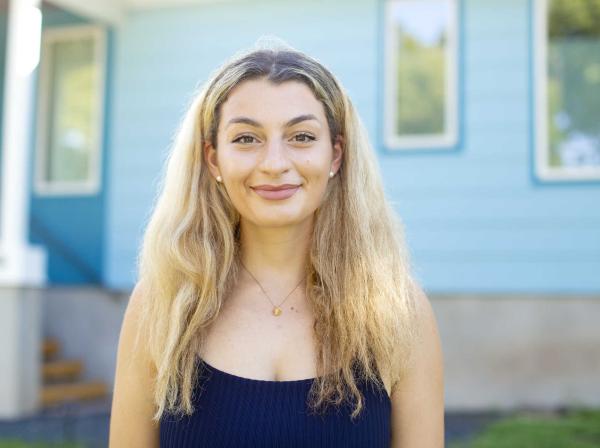 The image shows a young woman with long blonde hair, smiling and looking directly at the camera. She is wearing a sleeveless navy blue top, pearl earrings, and a simple pendant necklace. The background features a light blue building with white trim and windows, suggesting an outdoor setting. The overall atmosphere is bright and cheerful, with natural lighting enhancing the image.