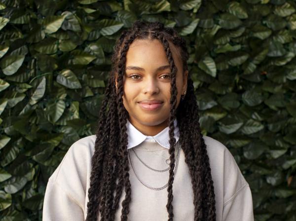 Taylor Huey wears a neutral tan sweatshirt with a white color shirt and multiple necklaces and stands in front of a green ivy background.