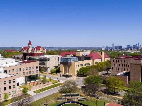 he image depicts an aerial view of a campus with various buildings. In the foreground, multi-story buildings with distinct architectural designs are visible, including one with a prominent red roof. The campus is well-maintained, surrounded by green spaces. The clear sky above and distant city skyline suggest an urban location. No people or vehicles are visible, creating a serene atmosphere. This image likely showcases the layout and proximity of an educational institution to the nearby city.