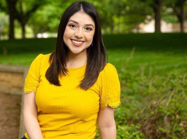 Victoria Garcia wears a yellow shirt and sits on a bench on Ragsdale patio. Greenery and trees are in the background.