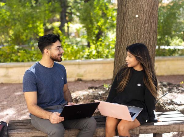  The image depicts two individuals sitting on a bench outdoors. Both are using laptops—one person wears a blue t-shirt, while the other wears a black long-sleeve shirt. They appear focused on their screens, suggesting work or study. The setting includes trees, creating a peaceful atmosphere.