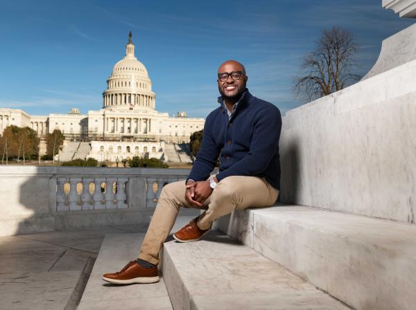 The image features a man sitting on marble steps with the United States Capitol building prominently in the background. He is smiling, wearing a dark blue cardigan, beige pants, and brown shoes. The setting is outdoors under a clear blue sky. The composition highlights both the individual and the iconic architecture, suggesting a professional or formal context. The overall atmosphere is bright and positive.