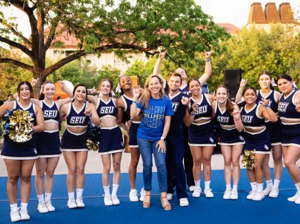 The image shows a group of cheerleaders and a few other individuals posing together outdoors. They are wearing matching navy blue and white uniforms with "SEU" printed on them. One person in the middle is wearing a blue T-shirt with "HILLFEST" written on it. Everyone is smiling and making a hand gesture, indicating a spirited and joyful atmosphere. The background features trees and a building, suggesting a campus or park setting.