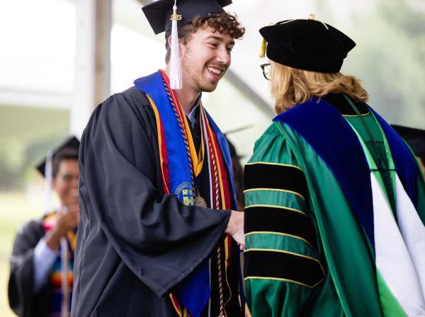 August Railey shakes hands with dean Catherine Campbell at commencement. Both are wearing caps and gowns for their commencement regalia.