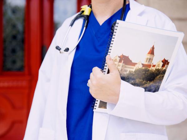 A person stands in front of hte red doors wearing blue scrubs a white coat and a stethoscope