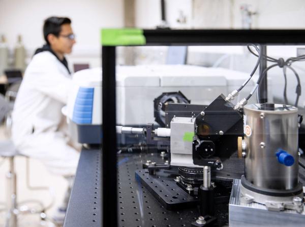 A student in a lab coat is in the background as a piece of research equipment is in focus in the foreground.