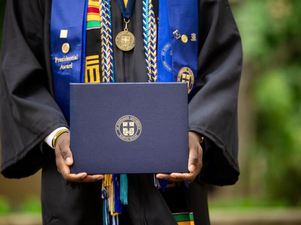 A student adorned with stoles and cords in their cap and gown holds their degree.