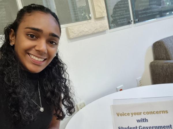 Analee Maharaj wears a black blouse and necklace and takes a selfie while sitting at a white table.