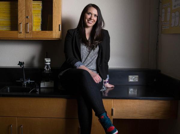 The image shows a woman sitting on a black countertop in a laboratory or classroom setting. She is smiling and casually dressed in a gray shirt, black blazer, and black pants with colorful sneakers. Behind her are wooden cabinets, a poster depicting animal and plant cells, and a microscope on the counter. The lighting is soft, with natural light coming from a window on the right, creating a relaxed and approachable atmosphere.