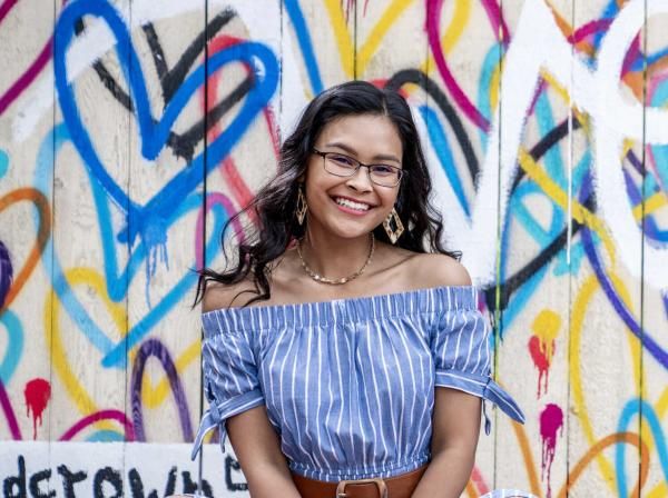 The image features a young woman with long, dark, wavy hair wearing glasses. She is smiling and dressed in an off-the-shoulder, blue and white striped top with short sleeves tied at the ends. She accessorizes with large, geometric earrings and a delicate necklace. The background is a vibrant mural with colorful heart shapes and abstract designs, adding an artistic and lively atmosphere to the image.