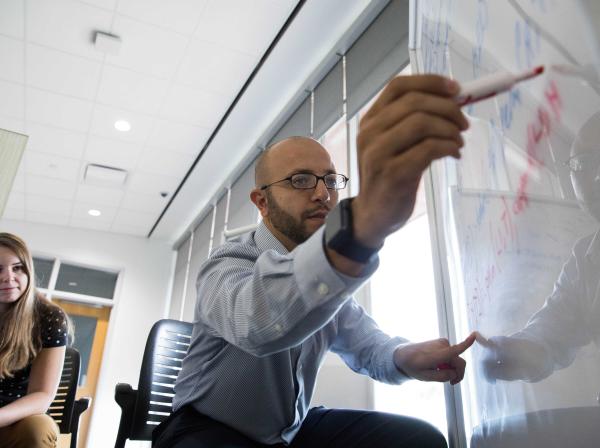 Professor writes on white board while student listens.