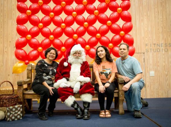 Santa Claus, a student and their family sit for a photo in front of a red balloon backdrop.