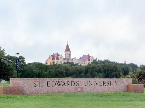 The entrance sign to St. Edward's University, with trees and Main Building in the distance.