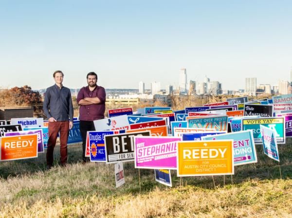 Jason Callahan and Omar Dominguez stand with a lot of candidate election signage with a view of the skyline in the background.