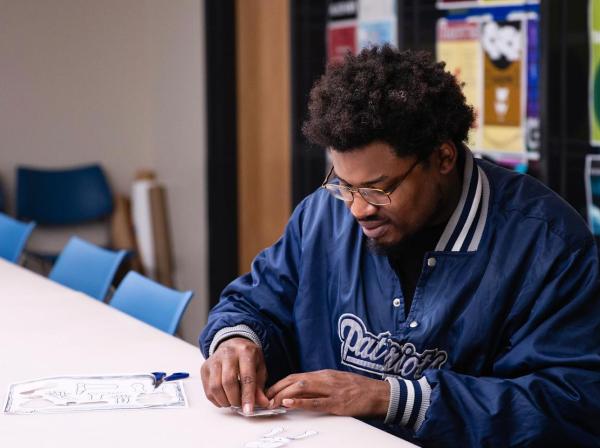 The image shows a man with glasses and a beard, wearing a blue "Patriots" jacket, sitting at a table and concentrating on a craft project involving paper cutouts. He is working alone in a room with blue chairs and colorful posters on the walls, indicating a creative or educational setting.