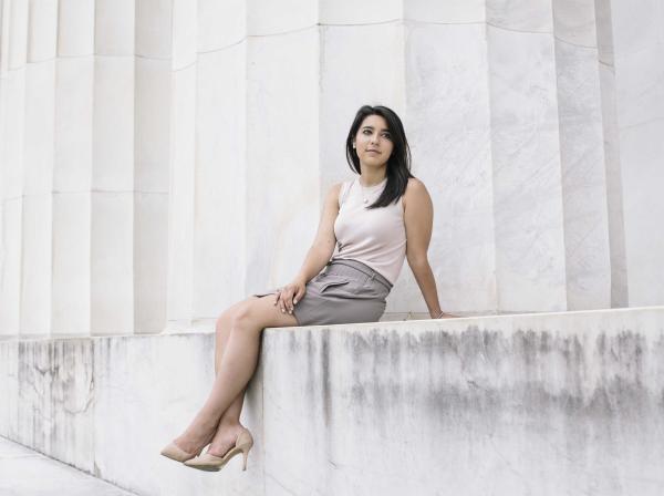 Victoria Ochoa wears a blouse, skirt and heels and sits on a ledge by massive columns.