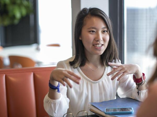 The image shows a young woman with shoulder-length hair, wearing a white blouse with a pearl neckline. She is sitting at a table in a casual setting, engaged in a conversation with another person. She has a blue wristband on her left wrist and a red watch on her right wrist. In front of her are a blue folder and a smartphone. The background includes a large window, some greenery, and blurred elements of a modern café or office space.