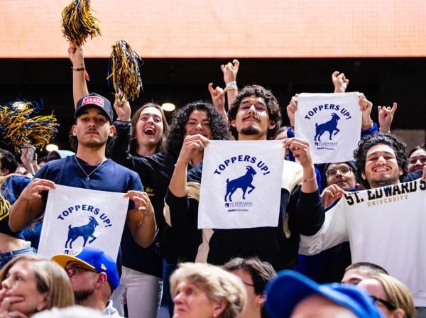 The image depicts a group of individuals at what appears to be a sporting event. They hold up signs or papers with the text "TOPPERS UP" and an image of a horse, suggesting they are fans of a sports team called the Toppers from St. Edwards University. The background includes an orange banner, likely part of the sports venue's decor. Overall, it captures the enthusiasm and community spirit of collegiate athletics.