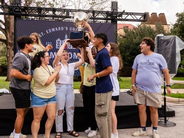 The image shows a group of people standing in front of a banner that reads "St. Edward's University." They are gathered around a trophy, which they are holding up together. Some of them are wearing t-shirts with the word "HILLTOPPERS" on them. It looks like a celebratory moment, possibly after winning a competition or event.