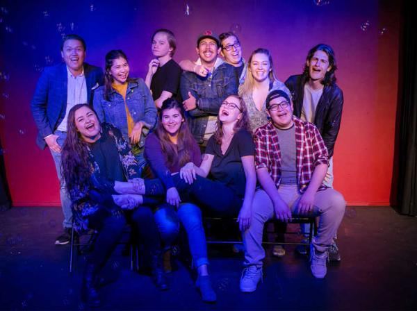 A group of improv students sit and stand and laughter together on a blue and, red and purple background as bubble float around them.