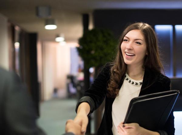 A student wearing a blazer holds a portfolio and shakes hand with their interviewer in an office.