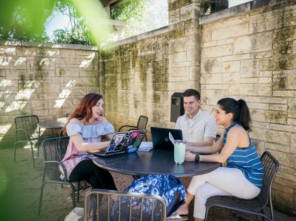 Three students sit at a patio table, hanging out, studying and working on their laptops.