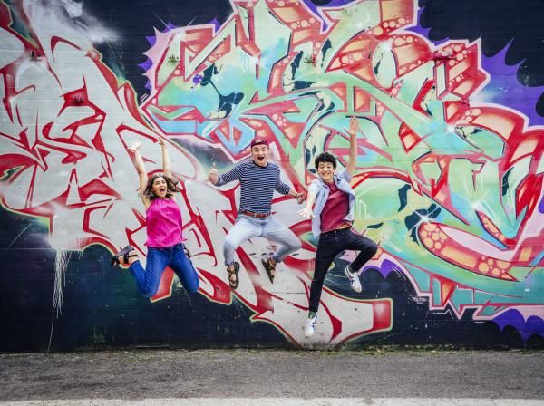 Three students jump in front of a colorful, patterned mural.