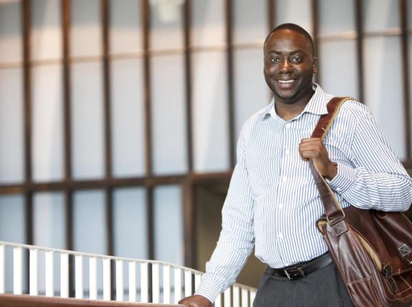 The image shows a smiling man wearing a light blue and white striped button-down shirt and gray trousers. He is carrying a brown leather shoulder bag. The background features a modern interior with large vertical wooden beams and frosted glass panels, giving a professional and contemporary ambiance. The man is positioned near a railing, suggesting he might be in an educational building.