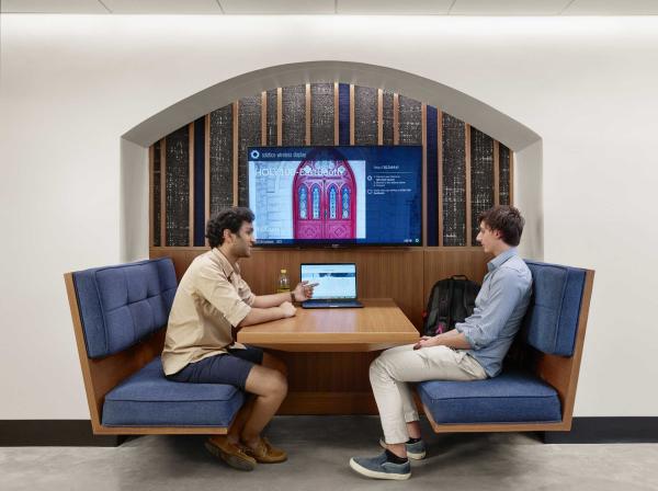 Two students sit in a booth with a table on the table as they look at the screen. A televsion mounted on the wall shows red doors.