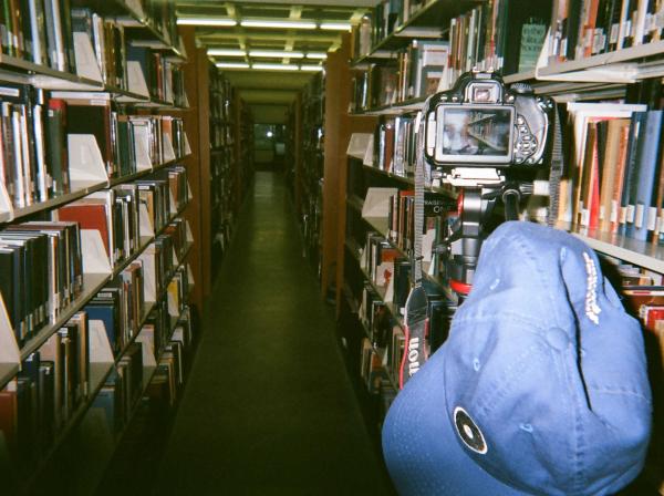 A camera and a hat on a tripod in between book stacks in the Munday Library.