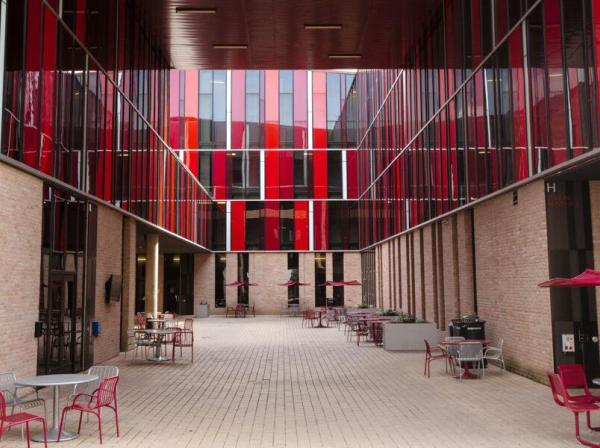 The image depicts a modern, open courtyard surrounded by buildings with striking red and glass facades. The buildings feature vertical red panels in varying shades, creating a dynamic and vibrant appearance. The courtyard is paved with light-colored bricks and is furnished with metal tables and chairs, some with red accents, and red umbrellas. The setting suggests a communal or educational environment, with a clean and contemporary architectural design.