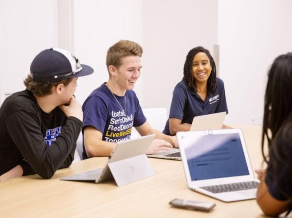 The image shows a group of four young adults sitting around a table, engaged in a lively discussion. They all have laptops in front of them and are smiling or laughing, indicating a positive and collaborative atmosphere. One person is wearing a baseball cap, another is in a branded T-shirt, and a third is wearing a polo shirt with a logo. The setting appears to be a casual meeting or study session in a bright, modern room, suggesting a school or university environment.