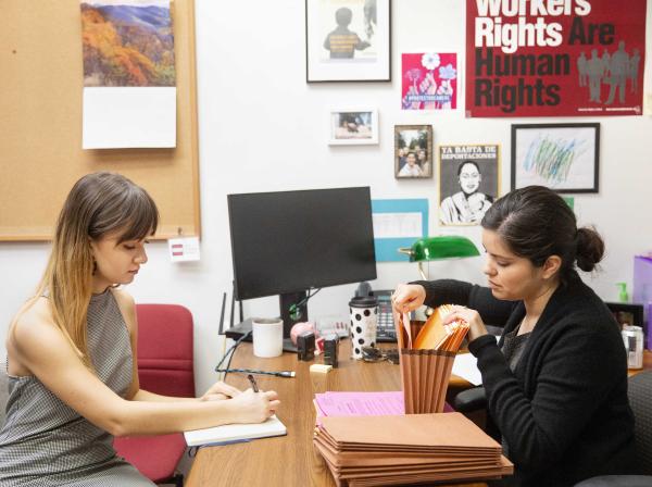 An intern and a staff member work together in an office at a desk, taking notes and filing paperwork.