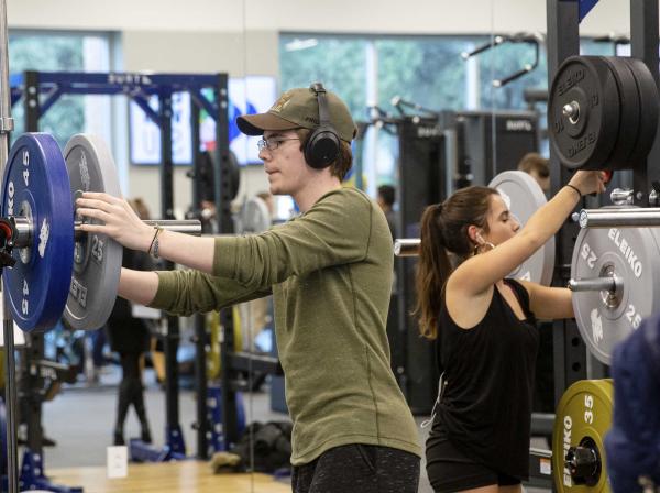 Two students use set up two different barbells on squat racks with weights.