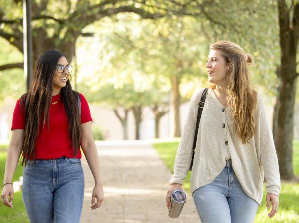 The image shows two young women walking outdoors on a tree-lined path, engaged in conversation. One woman, on the left, is wearing a red T-shirt, jeans, and glasses, while the other, on the right, is dressed in a light-colored cardigan, jeans, and is carrying a reusable tumbler. The scene is bright and sunny, with lush green trees in the background, suggesting a pleasant, casual walk, possibly on a campus or park. The overall mood is friendly and relaxed.