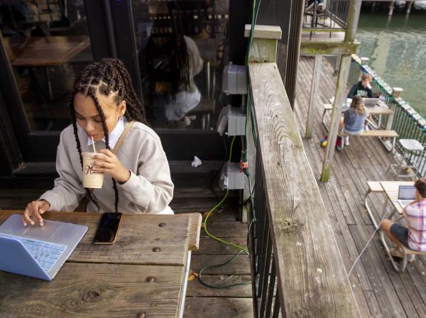 A student sits at a picnic table and drinks coffee while working on their laptop. Other people sitting at tables working on their laptops are seen below.