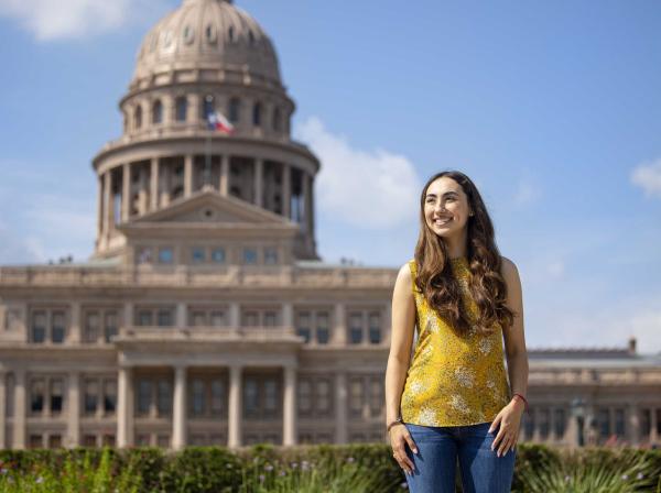 Michelle Flores stands outside of the Texas State Capitol wearing a yellow top and blue jeans on a sunny day.