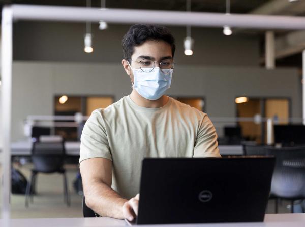 A student wears a face mask and types on a laptop in the Munday Library.