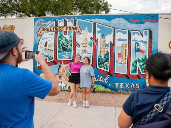 Two students pose in front of the Greetings from Austin mural as two people take their photo in the foreground.