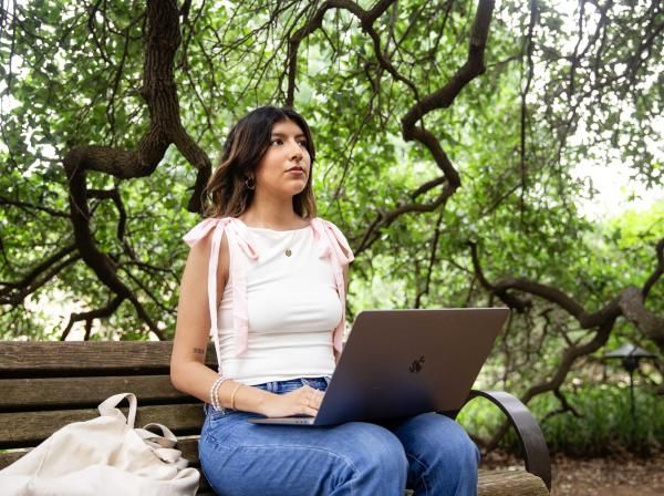 This image features a young woman sitting on a wooden bench in a serene, leafy outdoor setting. She is working on a laptop, with a focused expression, surrounded by twisted branches and green foliage. She wears a white top with pink shoulder ties, blue jeans, and minimal jewelry, including a pendant necklace and bracelets. A light-colored tote bag rests beside her on the bench.