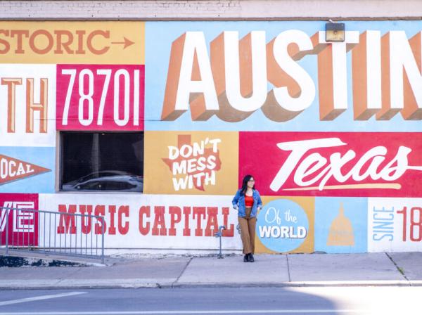 The image shows a large, colorful mural on the side of a building in Austin, Texas. The mural prominently features the words "Austin," "Texas," "Music Capital of the World," and "Don't Mess With Texas," along with other text and graphic elements. A woman is standing in front of the mural, casually posing with one hand on her hip and the other on the wall. She is wearing a denim jacket, red top, and brown pants. The scene captures a vibrant, urban atmosphere.